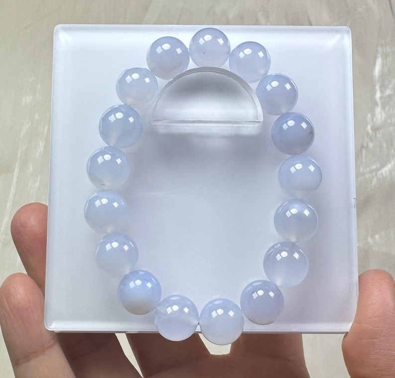 May include: A light blue beaded bracelet with round, polished beads. The bracelet is displayed on a white, rectangular stand with a clear, curved support. The beads have a translucent quality, and the bracelet is held by a hand.