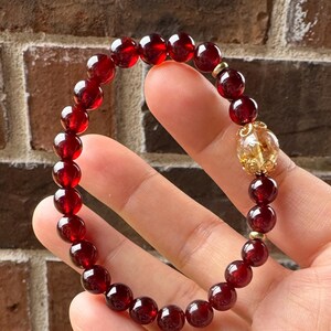 May include: A beaded bracelet featuring deep red garnet stones and a gold-colored accent bead with a clear, yellow gemstone. The bracelet is strung on an elastic band. The bracelet is shown on a hand.