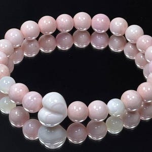 May include: A pink gemstone bracelet with a heart-shaped bead. The bracelet is made of round, polished beads in varying shades of pink and white. The bracelet is displayed on a reflective black surface.