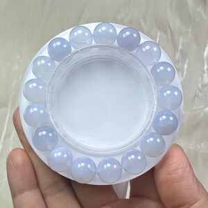 May include: A light blue beaded bracelet. The bracelet is made of round, translucent beads. The beads are arranged in a circular pattern around a white, circular container. The bracelet is displayed on a white surface.