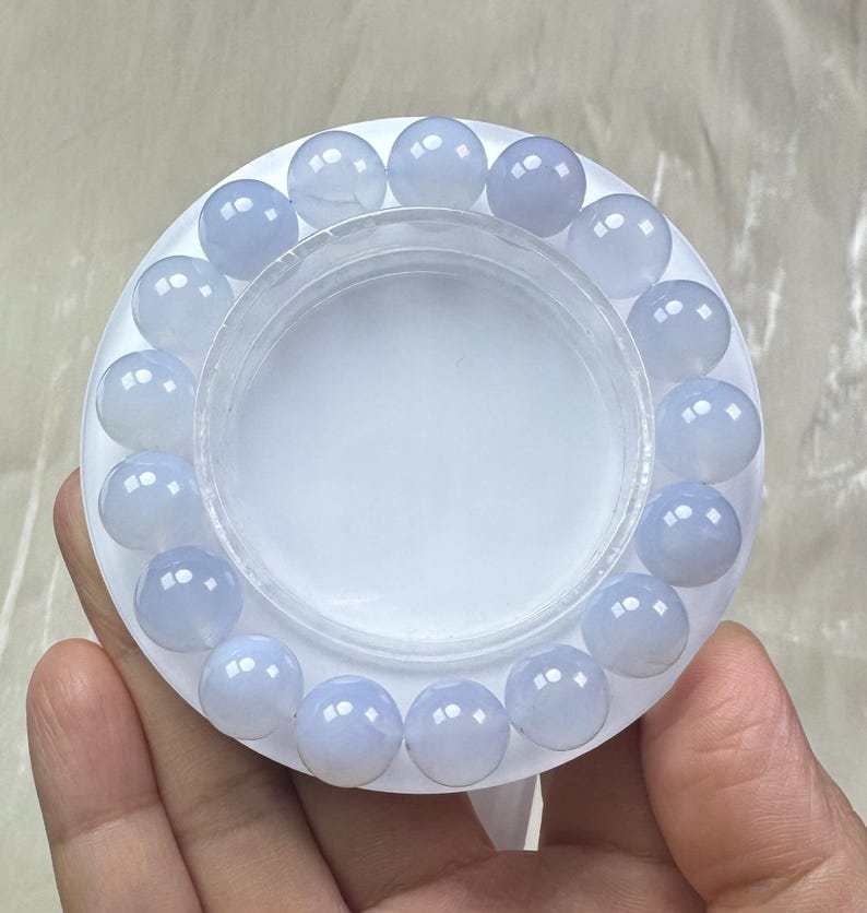 May include: A light blue beaded bracelet. The bracelet is made of round, translucent beads. The beads are arranged in a circular pattern around a white, circular container. The bracelet is displayed on a white surface.