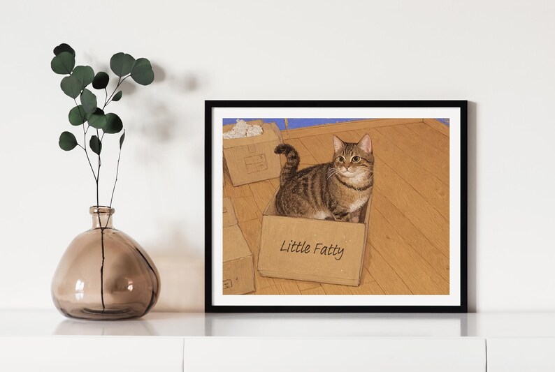 May include: A tabby cat sits inside a cardboard box with the words "Little Fatty" written on the side. The cat is looking directly at the viewer. The box is sitting on a wooden floor.
