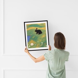 May include: A framed illustration of a black rabbit sitting in a field of green grass with yellow flowers and a small blue pond. The rabbit is looking to the right.