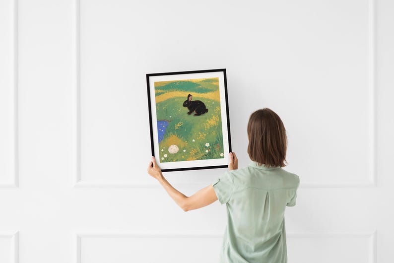 May include: A framed illustration of a black rabbit sitting in a field of green grass with yellow flowers and a small blue pond. The rabbit is looking to the right.