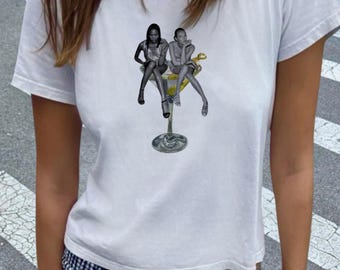 May include: White t-shirt with a graphic of two figures seated in a martini glass. The figures are in monochrome, and the martini glass has a yellow liquid and a lemon garnish. The t-shirt is a crew neck style.