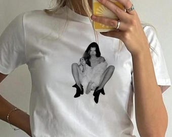 May include: White t-shirt featuring a black and white image of a woman sitting with her legs crossed, wearing a fur coat and boots.