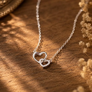 May include: A silver-toned necklace featuring two interlocking heart-shaped pendants. One heart is plain polished metal, while the smaller heart is encrusted with tiny clear stones. The delicate chain connects to both sides of the heart design.