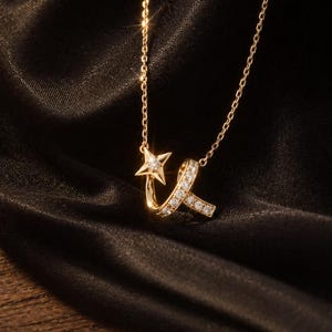 May include: A gold-colored necklace with a star pendant and a ribbon-like design adorned with small, sparkling stones. The necklace is displayed on a dark, textured background, highlighting its delicate details and elegant design.