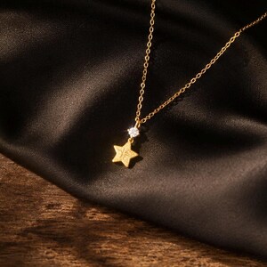 Può includere: Una collana color oro con una catenina sottile e un ciondolo composto da una piccola pietra preziosa trasparente a taglio rotondo posizionata sopra una stella a cinque punte testurizzata. Il metallo ha una finitura opaca.