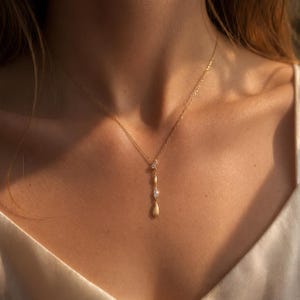 Könnte beinhalten: Eine zarte goldene Halskette mit einem Anhänger mit drei tropfenförmigen Elementen. Das obere Element hat einen kleinen, klaren Stein, das mittlere eine goldene Perle und das untere einen größeren, klaren Stein. Die Halskette wird am Hals getragen.