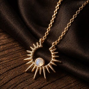 May include: A gold-coloured necklace featuring a sunburst pendant with a central, round, iridescent stone. The pendant has pointed rays extending outwards. The necklace chain is delicate and gold, resting on a dark brown fabric.