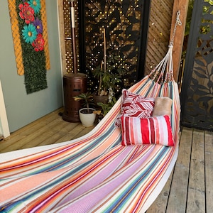 Puede incluir: Una hamaca de rayas multicolores con tejido de tonos vibrantes en naranja, rosa, azul y blanco. La hamaca incluye dos cojines decorativos, uno con un patrón de hojas en rojo y blanco y otro con rayas rojas y blancas.