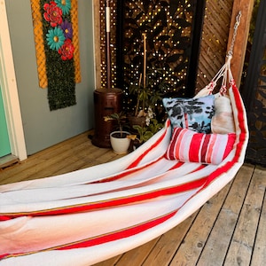 May include: A fabric hammock featuring wide white and red stripes with thin yellow and black accent lines. The hammock is suspended by metal chains and includes two rectangular throw pillows, one with a palm tree print and one with red stripes.
