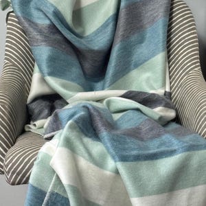Peut inclure: Une couverture douce à rayures dans les tons de bleu, de gris et de blanc est drapée sur une chaise. La couverture présente de larges rayures horizontales de largeurs et de tons variés, créant une esthétique chaleureuse et accueillante. La chaise a un motif rayé.
