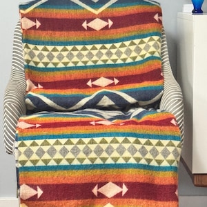 Könnte beinhalten: Eine farbenfrohe gewebte Decke mit geometrischem Muster liegt über einem gestreiften Sessel. Die Decke hat horizontale Streifen in Rot-, Orange-, Gelb-, Türkis- und Blautönen mit Diamant- und Pfeil-Designs in Weiß und Grau.