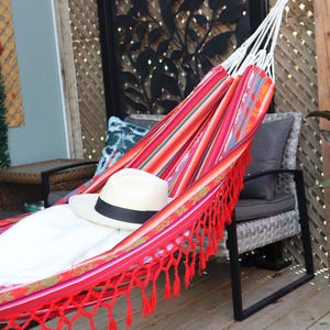 May include: A red and white striped hammock with red fringe hanging between two chairs. A white straw hat with a black band is lying in the hammock.