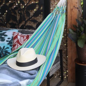 May include: A striped hammock with a white straw hat resting on top. The hammock is hanging from a wooden post and is surrounded by green plants.