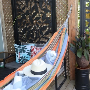 May include: A colourful striped hammock with a white hat and a blue blanket hanging from a metal hook on a wooden post. The hammock is hanging in front of a black metal lattice with a green plant in a brown pot to the right.