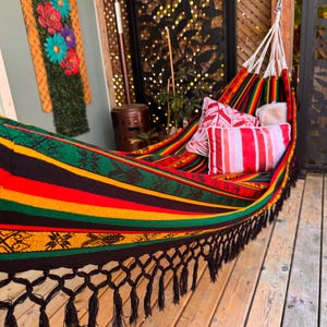 May include: A woven hammock featuring bold horizontal stripes in red, yellow, green, and black with intricate geometric patterns. The hammock includes black fringe along the bottom edge and two rectangular pillows with red and white striped designs.