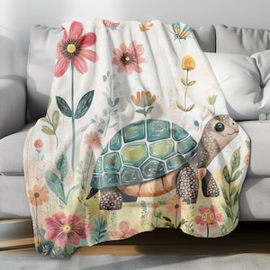 Puede incluir: Una suave manta blanca con un diseño de acuarela de una tortuga caminando por un campo de flores rosas, amarillas y verdes.