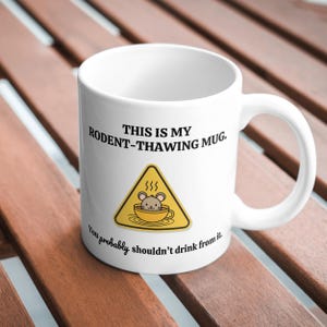 May include: White ceramic mug with a black warning sign that says "This is my rodent-thawing mug. You probably shouldn't drink from it." The sign has a yellow triangle with a black border and a cartoon mouse inside a mug.