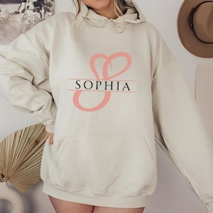 May include: A cream-coloured hoodie with a pink cursive letter "S" and the name "Sophia" printed on the front.