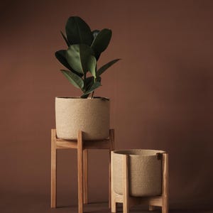 Może przedstawiać: Dwie plecione donice z juty, jedna z rośliną o dużych liściach, stoją na drewnianych stojakach na brązowym tle. Większa donica jest podniesiona na wysokim stojaku, a mniejsza spoczywa na krótszym.