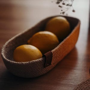 May include: A woven basket filled with three ripe lemons. The basket is oval-shaped and has a natural, textured appearance. A small leather tag is attached to the side. The lemons are bright yellow and sit on a wooden surface.