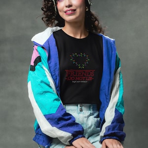 May include: A black t-shirt with the text "FRIENDS DO NOT LIE" and a heart-shaped design made of colourful lights. The shirt is worn with a blue, white, and pink windbreaker and denim shorts. The overall theme is a retro, 80s-inspired aesthetic.