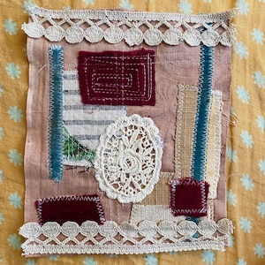 May include: A textile art piece with a pink background, featuring various fabric patches in burgundy, beige, and blue. A white lace oval and scalloped trim add detail. The piece is hand-stitched, showcasing a unique, handcrafted aesthetic.