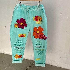 May include: Light turquoise trousers with an elastic drawstring waist and rolled cuffs. The trousers feature various fabric patches including large magenta, orange, and yellow flowers, a yellow teapot, and horizontal strips of green patterned fabric.