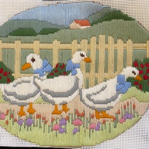 May include: A hand-stitched needlepoint scene depicting three geese wearing blue neck scarves, walking past a wooden fence. The background features rolling green hills, a small red-roofed house, and colourful flowers.