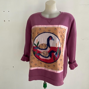May include: A mauve long-sleeve sweatshirt featuring a central rectangular panel with a floral background. The panel contains a circular applique depicting two geese in blue, white, and patterned fabric, surrounded by green cattails and red accents.