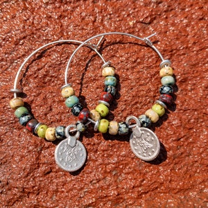 May include: A pair of silver hoop earrings with colorful ceramic beads and silver coin charms. The beads are green, blue, red, black, and white.