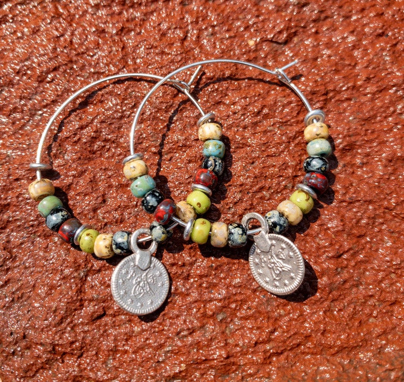 May include: A pair of silver hoop earrings with colorful ceramic beads and silver coin charms. The beads are green, blue, red, black, and white.
