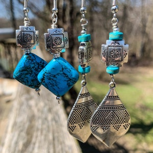 May include: A pair of silver dangle earrings with turquoise stones and geometric designs. The earrings feature a silver cross with a turquoise stone in the center, followed by a turquoise stone in a square shape, and then a silver teardrop-shaped pendant with geometric designs.