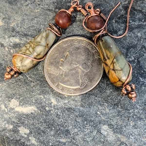 May include: A pair of copper wire wrapped earrings with brown and green stone beads. The earrings are shown on a gray stone surface with a US quarter for scale.