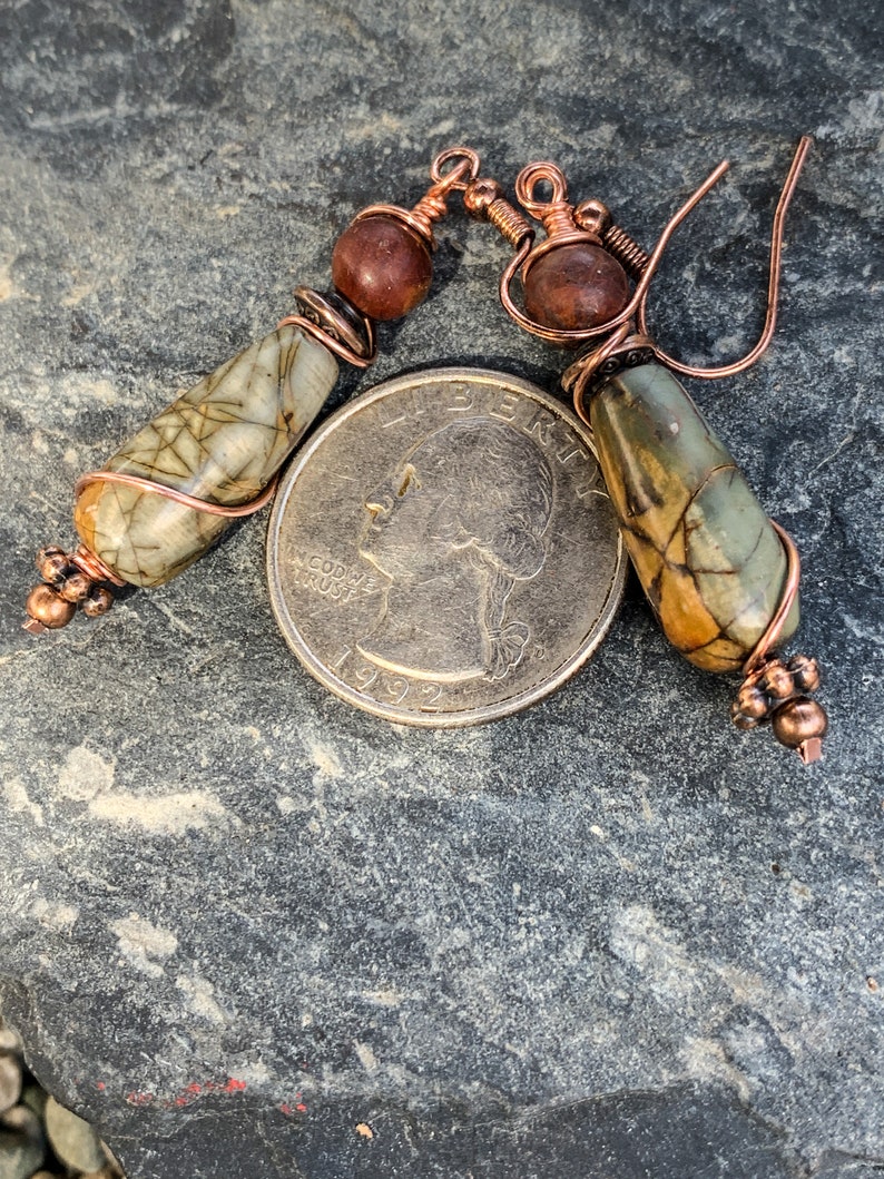 May include: A pair of copper wire wrapped earrings with brown and green stone beads. The earrings are shown on a gray stone surface with a US quarter for scale.