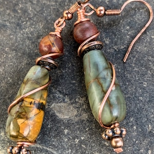 May include: A pair of copper wire wrapped earrings with green and brown stone beads. The earrings have a teardrop shape and are accented with copper wire.