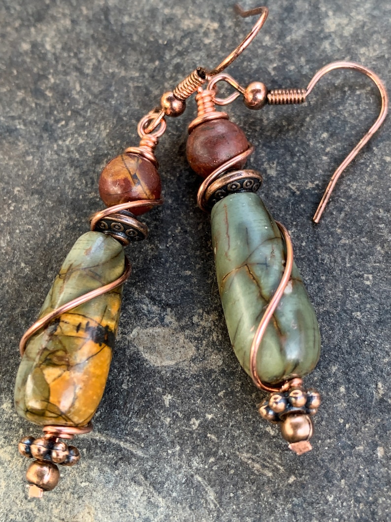 May include: A pair of copper wire wrapped earrings with green and brown stone beads. The earrings have a teardrop shape and are accented with copper wire.