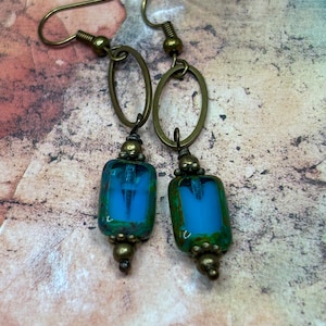 May include: A pair of dangle earrings featuring rectangular blue glass beads with mottled green edges. Each earring has a bronze-toned metal oval hoop at the top, connected to the bead by small bronze-toned metal spacer beads and a hook.