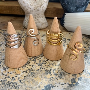 May include: Four rings displayed on wooden cone-shaped stands. The rings are silver and gold, featuring various designs, including wavy and spiral patterns. The rings are displayed on a light-colored surface.