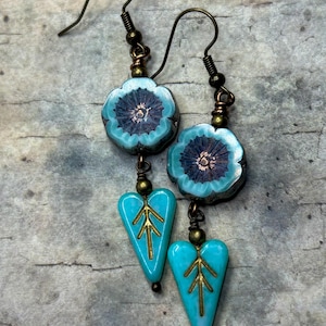 May include: A pair of drop earrings featuring a turquoise blue flower bead with a dark center, suspended above a turquoise blue heart-shaped bead with a gold-colored branch design. The earrings are attached to bronze-toned metal ear wires.