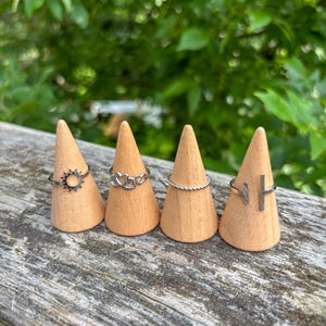 May include: Four silver rings displayed on wooden cone-shaped stands. The rings feature various designs, including a sunburst, hearts, a twisted band, and a minimalist bar. The stands are arranged on a weathered wooden surface.