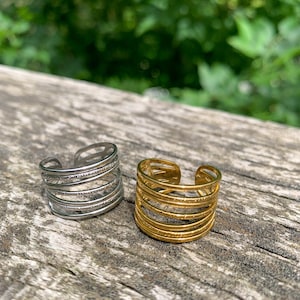 May include: Two open-ended rings, one silver and one gold, sit on a weathered wooden surface. The rings feature a multi-layered design with horizontal bands. The background is a blurred view of green foliage.