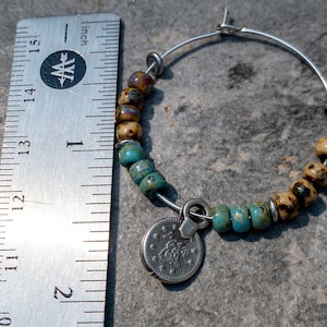 May include: A silver hoop earring with a charm attached. The charm is a silver coin with a design on it. The hoop is decorated with turquoise and brown beads.