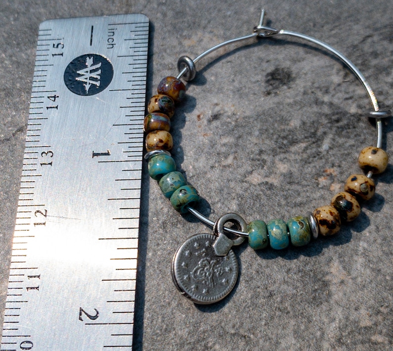 May include: A silver hoop earring with a charm attached. The charm is a silver coin with a design on it. The hoop is decorated with turquoise and brown beads.