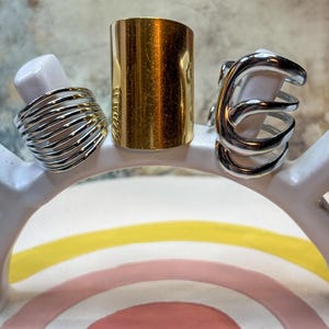 May include: Three rings displayed on a white, rainbow-shaped stand. The rings include a silver wire-wrapped ring, a gold-colored cylinder ring, and a silver cage-style ring. The stand features a pink, yellow, and pink rainbow design.
