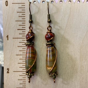 May include: A pair of dangle earrings with brown and red stone beads wrapped in copper wire. The earrings have a drop shape and are approximately 2 inches long.