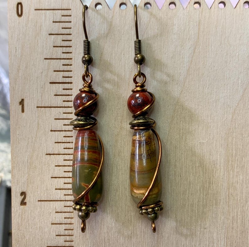 May include: A pair of dangle earrings with brown and red stone beads wrapped in copper wire. The earrings have a drop shape and are approximately 2 inches long.